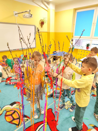 grupa przedszkolaków bawiąca się na kolorowym dywanie trzymająca kolorowe wstążki i ubierająca drzewo