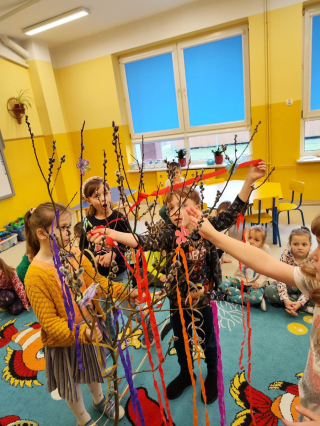 grupa przedszkolaków bawiąca się na kolorowym dywanie trzymająca kolorowe wstążki i ubierająca drzewo