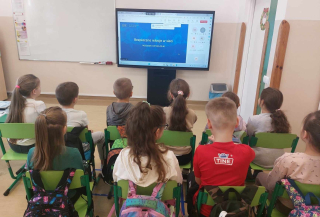 Grupa uczniów siedząca na krzesełkach i oglądająca weninar Bezpieczne relacje w sieci na monitorze interaktywnym