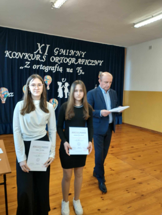 Dyrektor wręczający nagrodę 2 uczennicom w konkursie na granatowym tle z napisem XI Gminny Konkurs Ortograficzny „Z ortografią na TY ”