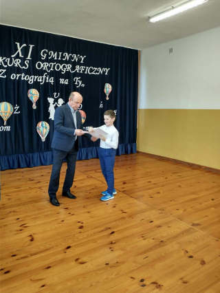 Dyrektor wręczający nagrodę uczniowi w konkursie na granatowym tle z napisem XI Gminny Konkurs Ortograficzny „Z ortografią na TY ”