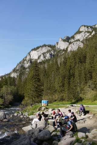 uczniowie siedzący na kamieniach nad potokiem, w tle góry