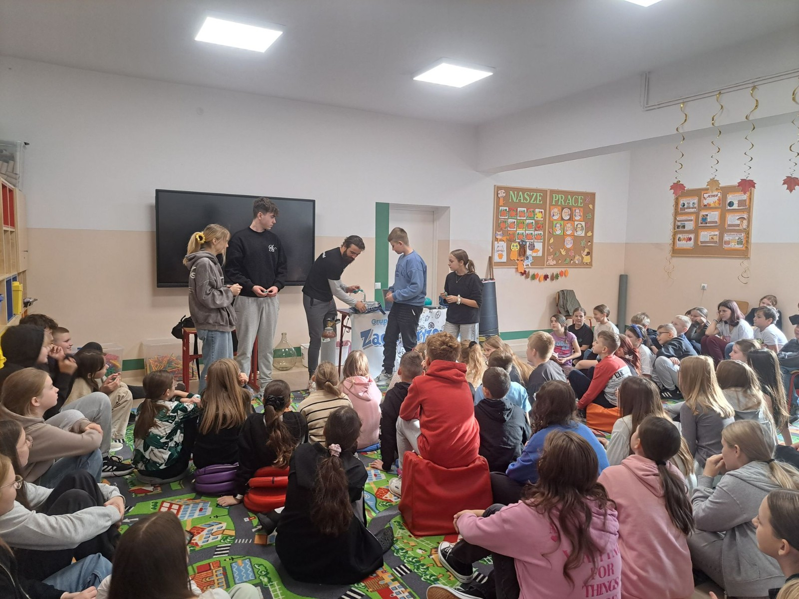 Zdjęcie przedstawiające dorosłych i młodzież podczas pokazu naukowego w klasie. Cztery osoby (prawdopodobnie prowadzący i asystenci) stoją przy stole z różnymi rekwizytami, prezentując coś publiczności. Liczna grupa dzieci i młodzieży siedzi na dywanie i obserwuje. Widać bannera z logo "Zae[le]ktryzowani".