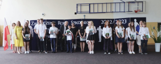 zdjęcie grupowe uczniów z wychowawcami, rodzicami i dyrekcją. W tle jasna ściana i kosz na hali sportowej, dekoracja na niebieskim tle z napisem zakończenie roku szkolnego oraz flagi Polski
