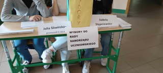 3 uczniów (członków komisji wyborczej do samorządu uczniowskiego) siedzących przy stoliku z napisami o ich funkcjach