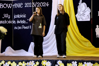 2 uczennice śpiewające na scenie, w tle dekoracja w kolorze granatowym z cytatem, flagą papieską oraz JP 2