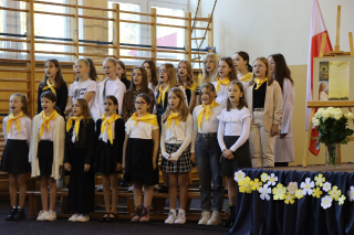 chór szkolny ubrany na galowo z żółtymi chustami na szyi, kwiaty stojące obok chóru w kolorach papieskich
