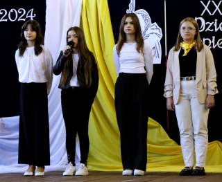 4 uczennice przemawiające do mikrofonu stojące na tle dekoracji granatowej i kolorów białym, żółtym flagi papieskiej
