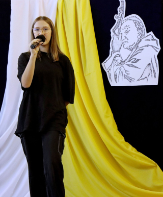 uczennica uśpiewająca na scenie, w tle dekoracja w kolorze granatowym z cytatem oraz JP 2